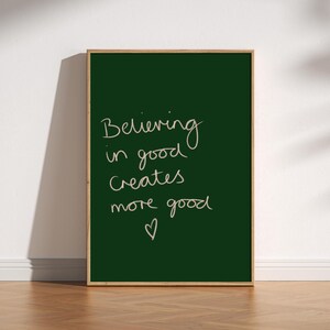 Peut inclure: Une affiche encadrée avec un fond vert foncé et le texte manuscrit "Believing in good creates more good" en beige clair. Un petit cœur est dessiné en dessous du texte. Le cadre est de couleur marron clair.