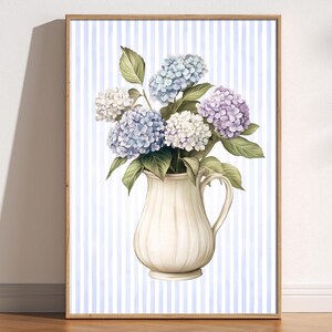 Peut inclure: Impression d'art encadrée avec une illustration à l'aquarelle d'hortensias dans une cruche crème. Les fleurs sont dans des tons de bleu, de violet et de blanc, avec des feuilles vertes. Le fond a des rayures verticales bleues et blanches.
