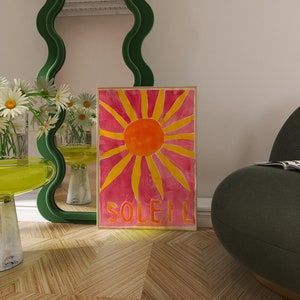 May include: A green wavy mirror with a pink and yellow sunburst print with the word "SOLEIL" at the bottom. The print is hanging on a wall next to a green armchair with a magazine on it.