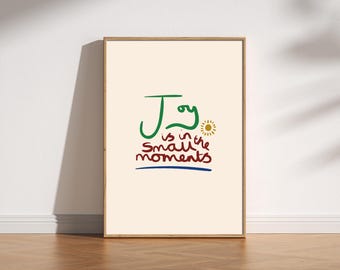 May include: Framed art print with the text "Joy is in the small moments" in green, red, and blue. The artwork features a sun illustration and is set against a cream background. The frame is a light brown color.