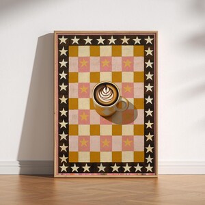 May include: A framed art print featuring a checkerboard design in pink, gold, and brown, adorned with stars. A cup of coffee with latte art sits atop the artwork, creating a visually appealing composition.