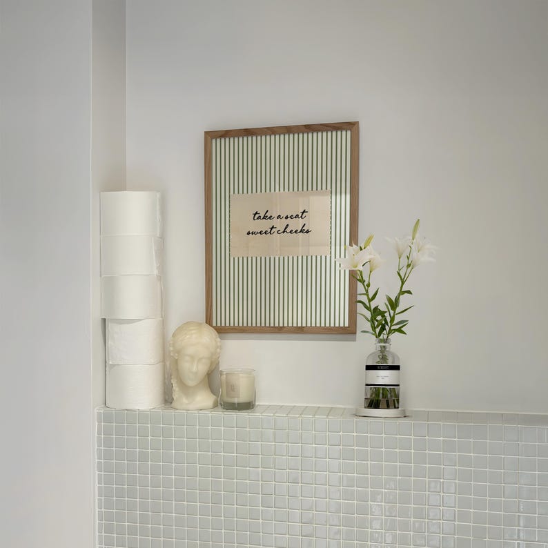 Funny bathroom wall art featuring olive green vertical pinstripes with a distressed cream scalloped label reading “take a seat sweet cheeks” in black handwritten script. Minimal vintage heritage inspired bathroom print with playful typography and a cosy boutique hotel aesthetic for guest bathrooms or powder rooms.