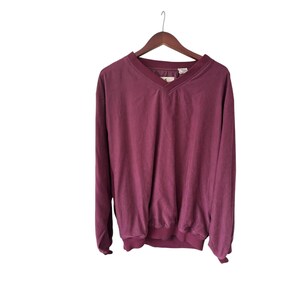 May include: A burgundy long-sleeved pullover shirt with a V-neck collar, hanging on a wooden hanger. The shirt has a ribbed hem and cuffs. The fabric appears to be a soft, slightly faded material.
