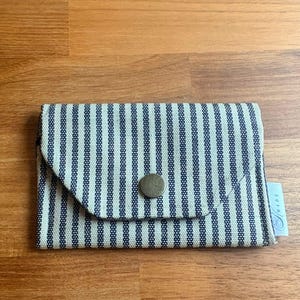 May include: A small, rectangular wallet with a blue and white striped pattern. The wallet has a flap closure secured by a round, bronze-coloured button. A small tag with cursive text is attached to the side. The wallet is made of fabric.