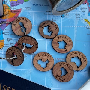 May include: A collection of circular wooden keychains featuring laser-cut silhouettes of various countries including Albania, Denmark, United States, France, Thailand, China, Australia, and Brazil, with the year 2023 engraved on the Brazil piece.