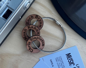 Puede incluir: Un llavero circular de cable metálico con tres anillos de madera grabados con los nombres Zach Bryan, Noah Kahan y Chris Stapleton. El llavero está colocado cerca de una entrada de concierto y una cámara, mostrando la textura de madera.