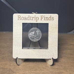 Peut inclure: Un cadre d'exposition en bois avec le texte "Roadtrip Finds" gravé en haut. Le cadre contient une pièce d'argent avec l'image d'un bâtiment et d'une personne. Le cadre est soutenu par deux petits supports en bois.