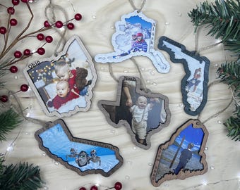 May include: Five wooden state-shaped Christmas ornaments featuring printed photographs and text. The ornaments are shaped like Ohio, Alaska, Florida, Texas, California, and Maine, each hanging from a jute twine loop and displaying personal family photos.