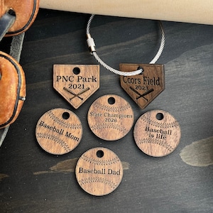 May include: A collection of six circular and home plate-shaped wooden tags with laser-engraved baseball designs and text including PNC Park 2021, Coors Field 2023, State Champions 2026, Baseball Mom, Baseball Dad, and Baseball is life, with a metal ring.