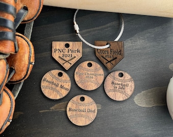 May include: A collection of six circular and home plate-shaped wooden tags with laser-engraved baseball designs and text including PNC Park 2021, Coors Field 2023, State Champions 2026, Baseball Mom, Baseball Dad, and Baseball is life, with a metal ring.