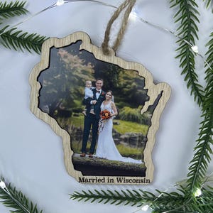 May include: A wooden ornament shaped like the state of Wisconsin featuring a printed photograph of a wedding couple and a child. The ornament has a light wood grain border and includes the text Married in Wisconsin at the bottom with a jute twine loop.