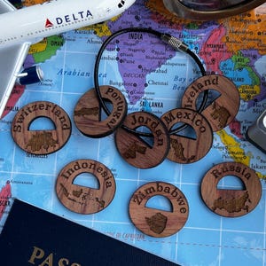May include: A collection of circular wooden keychains featuring engraved country names and map outlines, including Portugal, Switzerland, Jordan, Mexico, Ireland, Indonesia, Zimbabwe, and Russia, all strung together on a black metal cable ring.