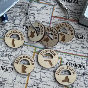 Könnte beinhalten: Eine Sammlung von sechs runden Holzschlüsselanhängern, die jeweils den Namen eines US-Bundesstaates und dessen geografischen Umriss lasergraviert zeigen. Enthalten sind Kalifornien, Texas, Alaska, Michigan, Minnesota, Florida und New York.
