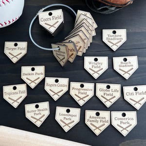 May include: A collection of light-coloured wooden baseball home plate tags, each engraved with a stadium name and crossed bats. The tags feature a hole at the top for attachment and are displayed with a metal cable ring and a baseball.