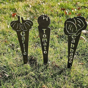 May include: Three metal garden markers with cut-out images of a tomato, garlic, and cabbage. The markers are painted black and have the names of the vegetables written on them.