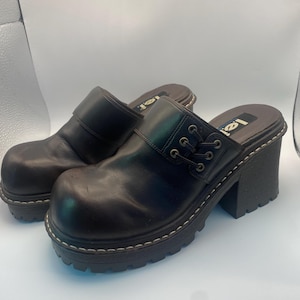 May include: A pair of black leather clogs with a chunky heel and a rounded toe. The clogs have a strap across the top with decorative lacing and a white stitched sole. The brand name "Lei" is visible on the insole.