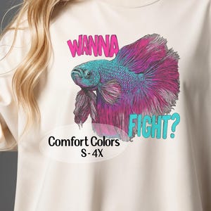 May include: Cream-colored t-shirt featuring a vibrant graphic of a betta fish with teal and magenta hues. The text "WANNA FIGHT?" is above the fish in pink and teal. The shirt also has the text "Comfort Colors S-4X".