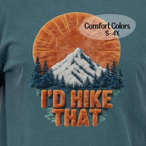 May include: Teal-colored t-shirt with a mountain and sun graphic. The graphic features a snow-capped mountain, orange sun, and the text "I'd Hike That" in orange. The shirt also has the text "Comfort Colors S-4X".