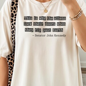 Op de afbeelding: Ivoor kleurig t-shirt met de quote "Dit is waarom de aliens hun deuren op slot doen als ze langs de aarde vliegen - Senator John Kennedy." Het woord "Ivory" staat onderaan het shirt.