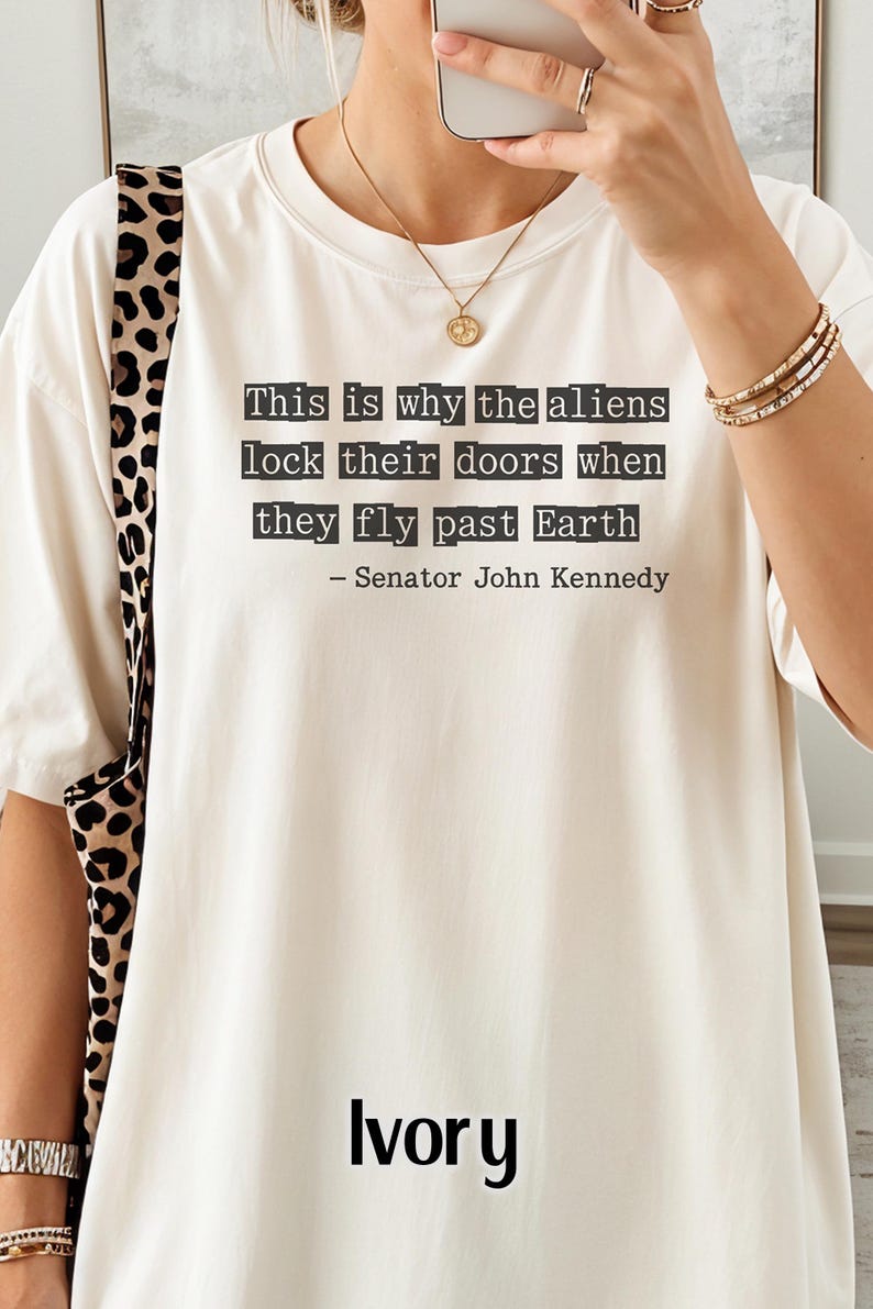Op de afbeelding: Ivoor kleurig t-shirt met de quote "Dit is waarom de aliens hun deuren op slot doen als ze langs de aarde vliegen - Senator John Kennedy." Het woord "Ivory" staat onderaan het shirt.