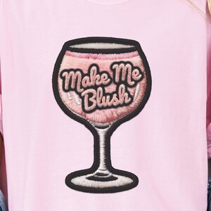 May include: Pink t-shirt featuring a wine glass design with the phrase "Make Me Blush" embroidered in a cursive font. The wine glass is outlined in black, with a pink interior. The shirt is a solid light pink color.