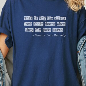 Op de afbeelding: Marineblauw T-shirt met de quote "Daarom sluiten de aliens hun deuren als ze langs de aarde vliegen - Senator John Kennedy" in witte tekst. Het shirt heeft een ronde hals.