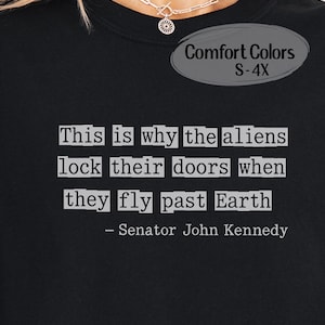 May include: Black Comfort Colors t-shirt with the quote "This is why the aliens lock their doors when they fly past Earth - Senator John Kennedy" in white text. The shirt is available in sizes S-4X.