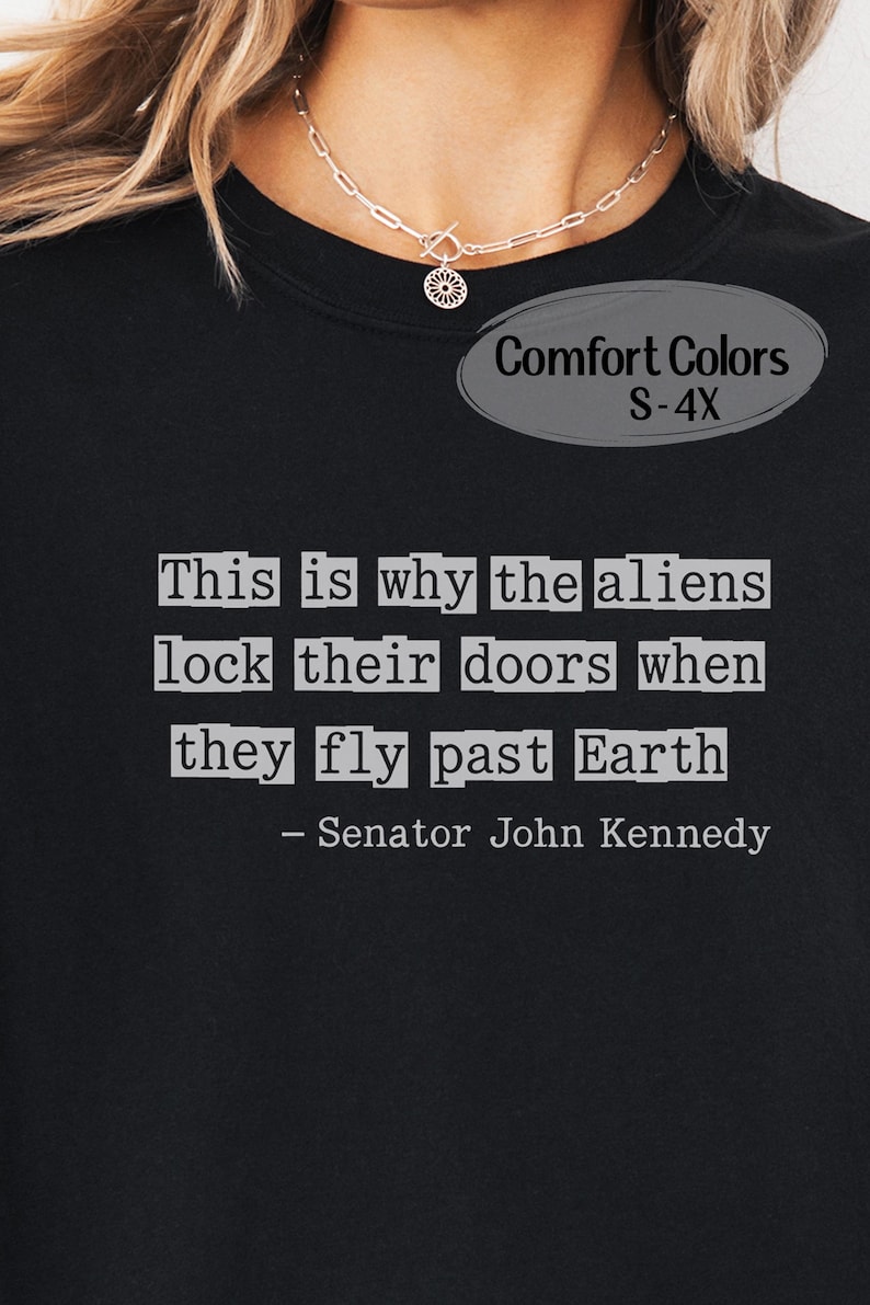 Op de afbeelding: Zwart Comfort Colors T-shirt met de quote "This is why the aliens lock their doors when they fly past Earth - Senator John Kennedy" in witte tekst. Verkrijgbaar in maten S-4X.