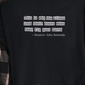 Op de afbeelding: Zwart sweatshirt met de tekst: "This is why the aliens lock their doors when they fly past Earth - Senator John Kennedy." Het woord "Black" staat in witte letters onderaan.