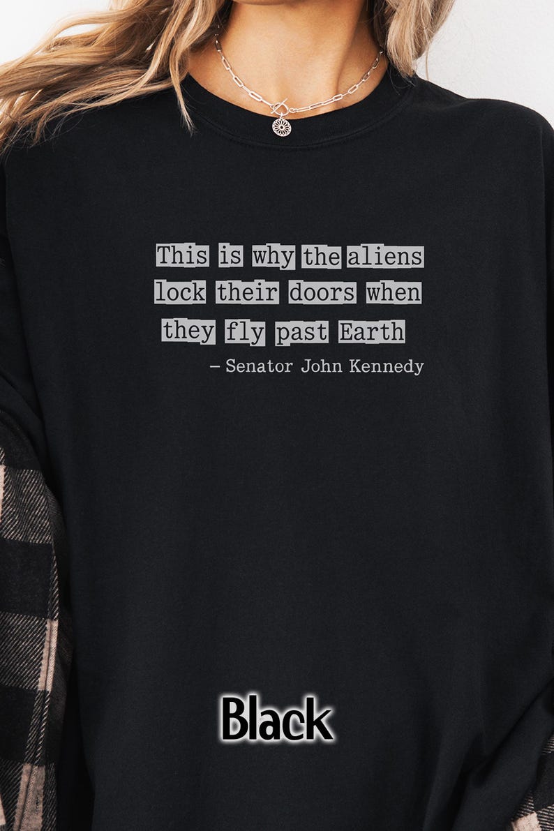 Op de afbeelding: Zwart sweatshirt met de tekst: "This is why the aliens lock their doors when they fly past Earth - Senator John Kennedy." Het woord "Black" staat in witte letters onderaan.