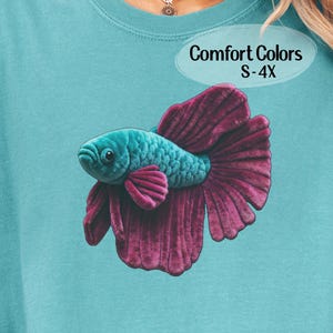 May include: A teal-colored sweatshirt featuring a detailed illustration of a betta fish. The fish has a turquoise body and a large, ruffled, magenta-colored tail. The text "Comfort Colors S-4X" is in an oval on the upper right.