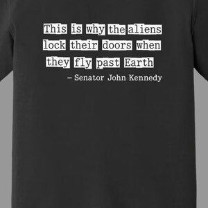 Op de afbeelding: Zwart t-shirt met witte tekst: "Dit is waarom de aliens hun deuren op slot doen als ze langs de aarde vliegen - Senator John Kennedy." Het woord "Zwart" staat onderaan gedrukt.