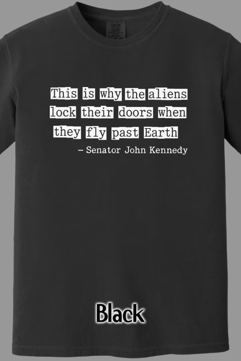 Op de afbeelding: Zwart t-shirt met witte tekst: "Dit is waarom de aliens hun deuren op slot doen als ze langs de aarde vliegen - Senator John Kennedy." Het woord "Zwart" staat onderaan gedrukt.