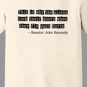 Op de afbeelding: Ivoor T-shirt met de tekst "This is why the aliens lock their doors when they fly past Earth - Senator John Kennedy" in zwarte letters. Het shirt heeft een ronde hals.