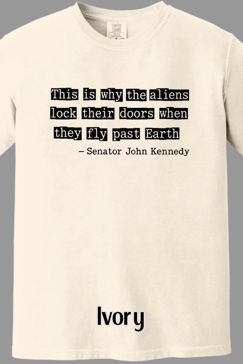 Op de afbeelding: Ivoor T-shirt met de tekst "This is why the aliens lock their doors when they fly past Earth - Senator John Kennedy" in zwarte letters. Het shirt heeft een ronde hals.