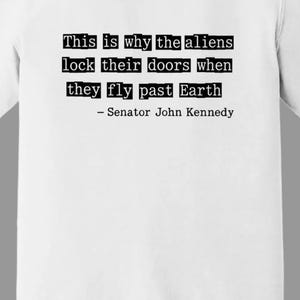Op de afbeelding: Wit T-shirt met de quote "This is why the aliens lock their doors when they fly past Earth - Senator John Kennedy" in zwarte letters. Het woord "White" staat in zwarte letters onderaan.