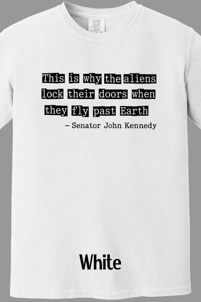 Op de afbeelding: Wit T-shirt met de quote "This is why the aliens lock their doors when they fly past Earth - Senator John Kennedy" in zwarte letters. Het woord "White" staat in zwarte letters onderaan.