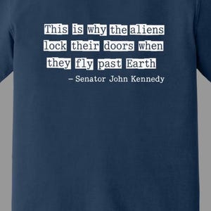 Op de afbeelding: Marineblauw T-shirt met witte tekst: "Dit is waarom de aliens hun deuren op slot doen als ze langs de aarde vliegen - Senator John Kennedy." Het shirt heeft een Comfort Colors-label.