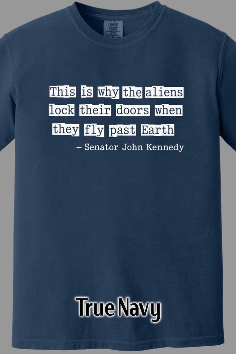 Op de afbeelding: Marineblauw T-shirt met witte tekst: "Dit is waarom de aliens hun deuren op slot doen als ze langs de aarde vliegen - Senator John Kennedy." Het shirt heeft een Comfort Colors-label.