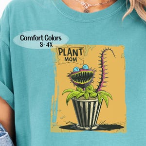 May include: Teal Comfort Colors t-shirt featuring a cartoon plant monster in a striped pot. The plant has a large mouth, blue eyes, and a spiky stem. The text "PLANT MOM" is above the plant, and "Comfort Colors S-4X" is above the text.