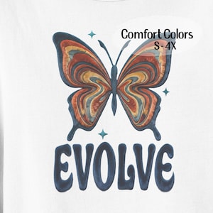 May include: White t-shirt featuring a colorful butterfly design with the word "EVOLVE" in navy blue. The butterfly has orange, yellow, and blue details. The text "Comfort Colors S-4X" is also visible.