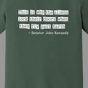 Op de afbeelding: Blauwspar T-shirt met de quote "This is why the aliens lock their doors when they fly past Earth - Senator John Kennedy" in witte tekst. Het shirt is effen van kleur.
