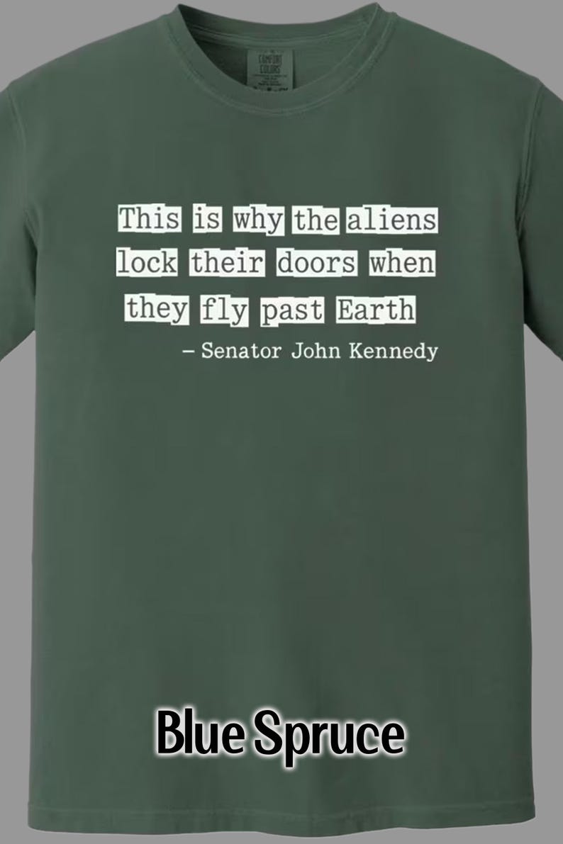 Op de afbeelding: Blauwspar T-shirt met de quote "This is why the aliens lock their doors when they fly past Earth - Senator John Kennedy" in witte tekst. Het shirt is effen van kleur.