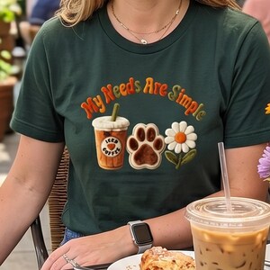 May include: Dark green t-shirt with the embroidered text "My Needs Are Simple." The design includes an iced coffee, a paw print, and a daisy. A clear plastic cup of iced coffee and a pastry are on a table.