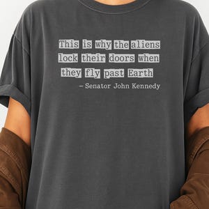 Op de afbeelding: Grafietkleurig T-shirt met de quote "This is why the aliens lock their doors when they fly past Earth" toegeschreven aan Senator John Kennedy.