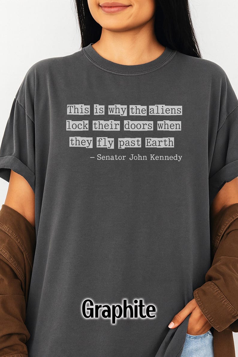 Op de afbeelding: Grafietkleurig T-shirt met de quote "This is why the aliens lock their doors when they fly past Earth" toegeschreven aan Senator John Kennedy.