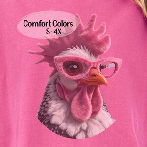 May include: Pink t-shirt featuring a whimsical illustration of a rooster wearing pink glasses. The rooster has a pink comb and wattle. The text "Comfort Colors S-4X" is in a white oval. The shirt is a solid pink color.