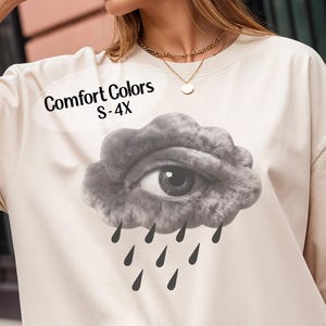 May include: Cream-colored Comfort Colors sweatshirt featuring a graphic of an eye within a gray cloud, with black raindrops falling below. The text "Comfort Colors S-4X" is displayed above the graphic. The sweatshirt has a relaxed fit.