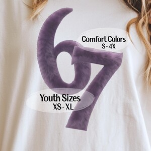May include: White t-shirt with a large, purple, fuzzy "67" graphic. Text on the shirt includes "Comfort Colors S-4X" and "Youth Sizes XS-XL". The shirt is a casual style.