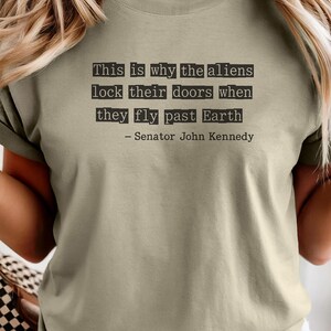 Op de afbeelding: Khaki-kleurig T-shirt met een citaat toegeschreven aan senator John Kennedy: "This is why the aliens lock their doors when they fly past Earth." Het citaat is in zwarte, typemachine-stijl lettertype. De kleur van het shirt is onderaan gelabeld met "Khaki".